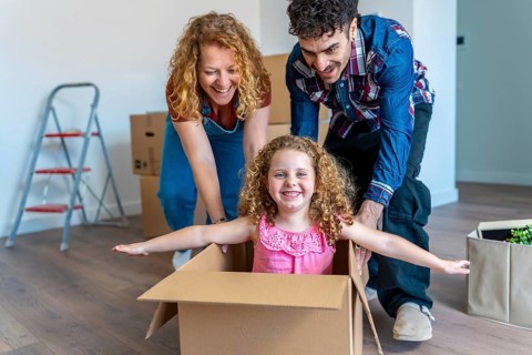 Parents pushing their young daughter in a cardboard box on a wooden floor, celebrating their
