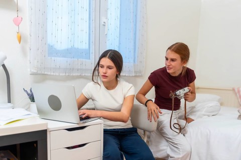 Teenage sisters sharing a bedroom, one focusing on studying or browsing online using a laptop while