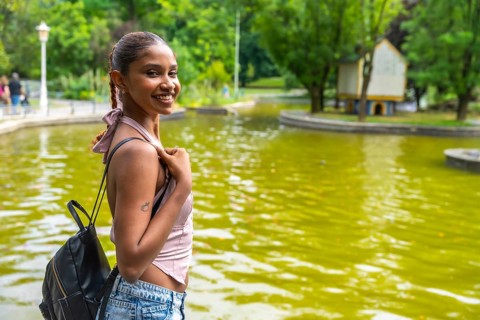Young woman with a backpack smiling, standing by a calm green pond in an urban park, enjoying a day