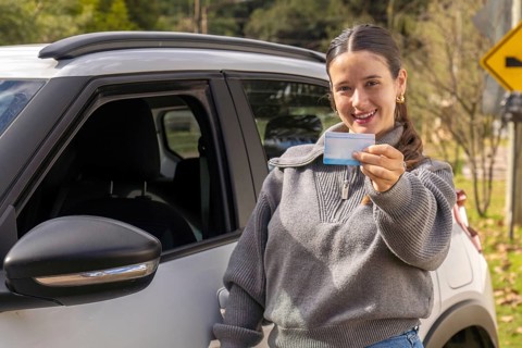 Young woman standing beside white car holding driving license, road curve warning sign visible in