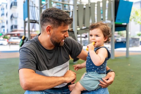 Father holding his infant son in a playground, the happy toddler enjoying a healthy snack during