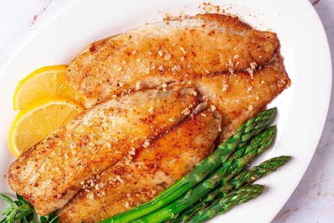Fresh fish filets are served on a white plate alongside green asparagus and slices of lemon. The