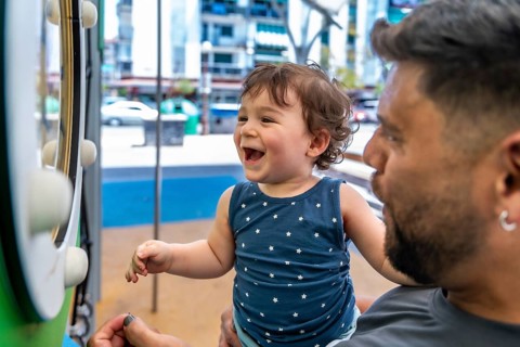 Toddler son laughing in father's arms during playful bonding at an urban park playground, candid