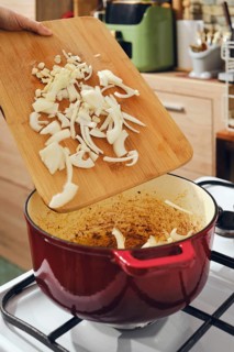 Person holds wooden cutting board with sliced onions above red cooking pot. Pot sits on stove with