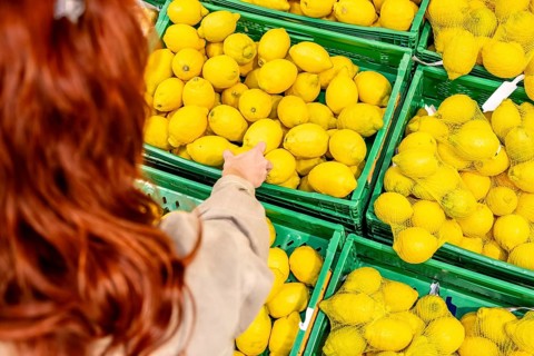 Redheaded woman selects bright yellow lemons from green crates at a supermarket produce display,