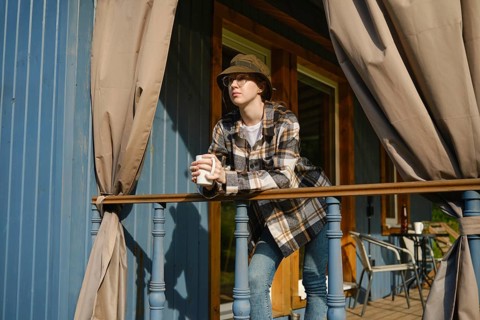 Young woman stands on terrace of wooden cabin leaning on railing holding cup of tea. She wears