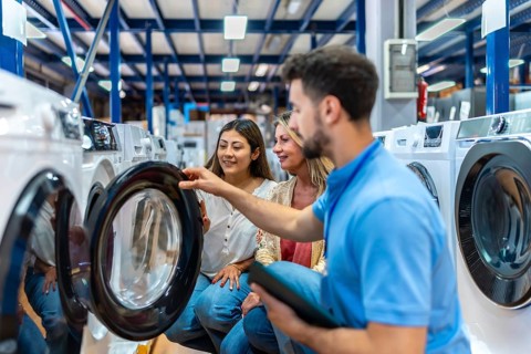 Salesperson assisting two women in a home appliance store, explaining features of a washing machine