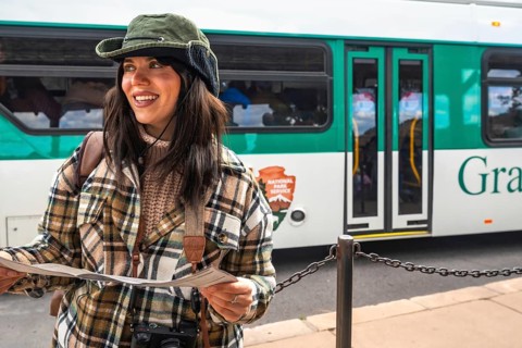 Smiling woman traveler in a plaid shirt and hat holds a park map at a shuttle bus stop, ready to