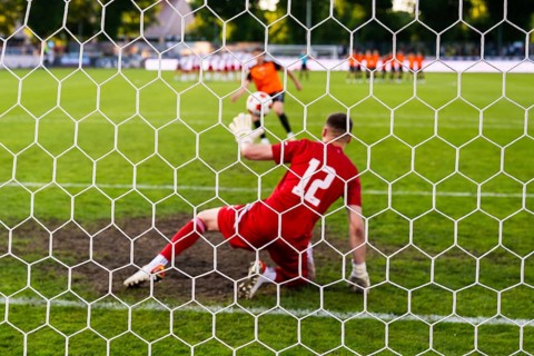 View through the net of a soccer goalkeeper in a red kit preparing to defend a penalty kick during