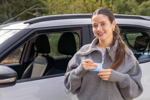 Smiling young woman standing beside car holding her driving license with both hands, outdoor