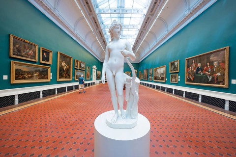 A white sculpture stands in a gallery corridor with red tiles flanked by paintings on green walls,