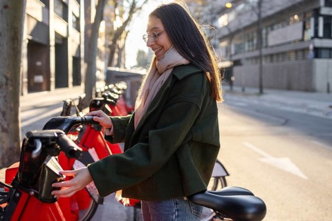 A smiling woman in a green coat rents a bicycle on a city street, glowing in the sunlight She