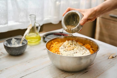 A person pours seasoning into a bowl filled with grated carrots and other ingredients. The kitchen