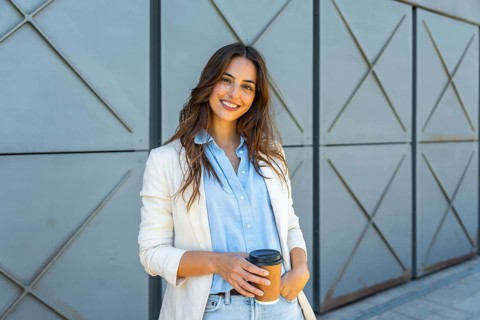 Confident young professional woman in a light blazer smiling outdoors, holding a reusable coffee