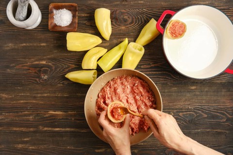 Hands are stuffing bell peppers with ground meat Nearby are halved peppers ready to be filled.