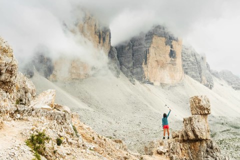 Female hiker raising trekking poles beneath misty Dolomites cliffs celebrating achievement