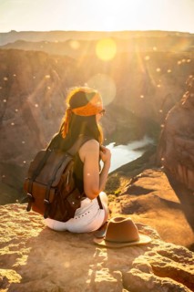 Woman traveler sitting with a backpack on a cliff edge, watching the panoramic sunset view over