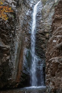 A high waterfall flows through a rocky gorge surrounded by autumn foliage and rough rocks,
