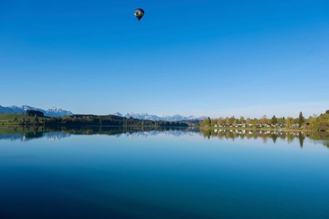 A hot air balloon floats over a lake with mountains in the background in a clear atmosphere, Lech