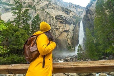 Woman wearing yellow rain jacket and beanie, carrying a backpack, standing on a wooden bridge