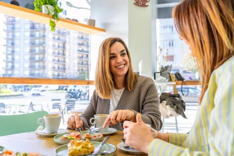 Two smiling women friends enjoying brunch and talking at a cafe table, with coffee and food,