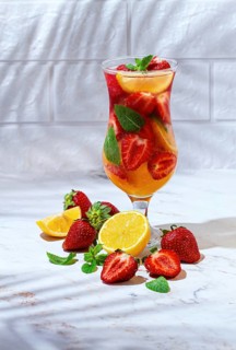 A tall glass filled with a colorful drink made from strawberries, lemon slices, and mint leaves.
