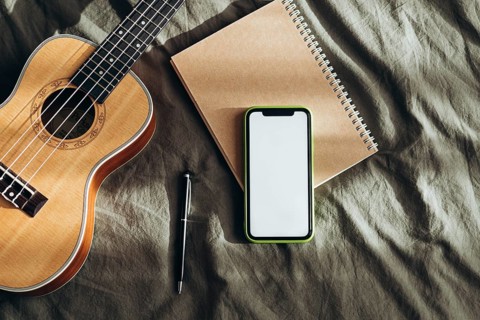 Cozy creative workspace featuring a ukulele, blank notebook, pen, and smart phone on a soft fabric