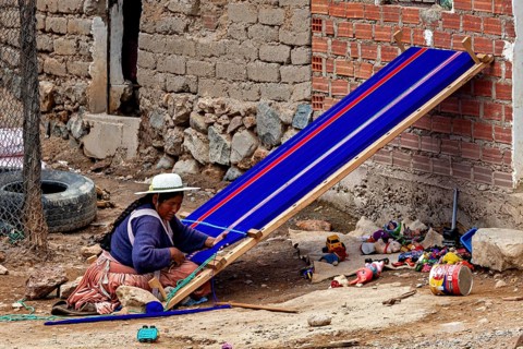 A woman weaves on a blue loom surrounded by brick walls and materials on a dusty courtyard, a woman