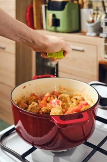 Hand is squeezing lime juice into pot filled with meat and vegetables on stove. Adding fresh