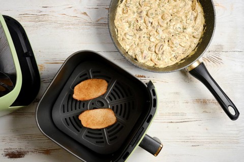 Two slices of meat are placed in air fryer basket. Skillet on tabletop contains creamy mushrooms.