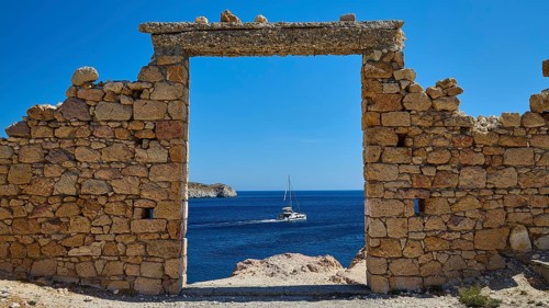 A stone arch with a view of a sailboat on the sea under clear blue sky, Palio Ergostasio Kaolini,