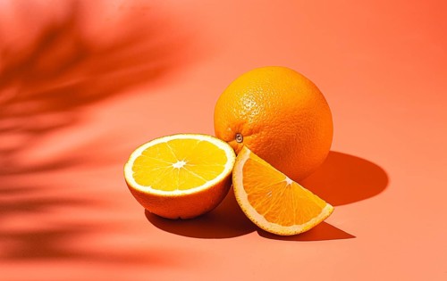 On the coral surface, next to two neatly sliced slices, there is a whole orange. Sunlight casts