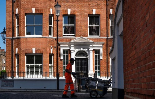 A street cleaner pushes a wheelbarrow with trash containers, in the background a red brick