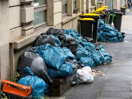 Mountains of trash made of blue and black bags pile up along a house façade on the sidewalk,