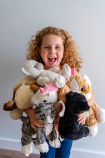 Little girl with curly hair laughing joyfully, holding a large pile of her beloved stuffed animals,