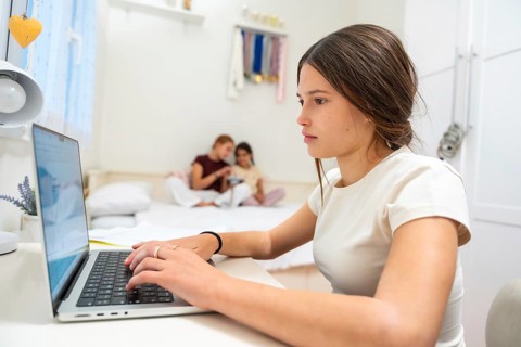 Young woman focused on her laptop doing homework while her sisters are relaxing on the bed in the