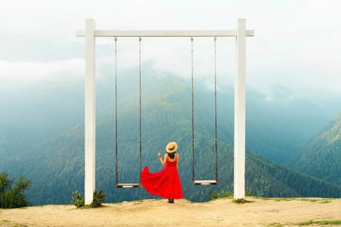 Woman wearing flowing red dress standing between mountain swings overlooking misty forest valley in