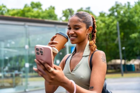 Happy young woman smiling while holding a reusable coffee cup and interacting with her cell phone