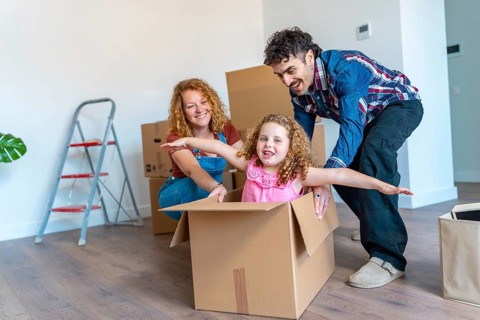 Happy family moving into a new home, parents pushing their smiling daughter inside a cardboard box,