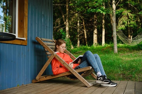 Girl sits on wooden chair on porch of wooden cabin reading book. Surrounding trees provide shade.