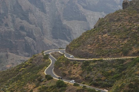 Steep serpentine road with cars snaking along a rock wall surrounded by vegetation, Masca, Barranco