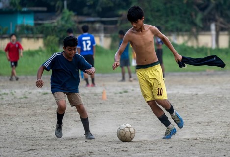 Children play football on a dusty neighbourhood field, chasing the ball with energy and