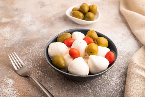 White Mozzarella cheese, with tomatoes and olives in blue bowl on brown concrete background and