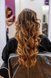 Woman's back with long, wavy balayage hair sitting in a salon chair while a hairdresser is working
