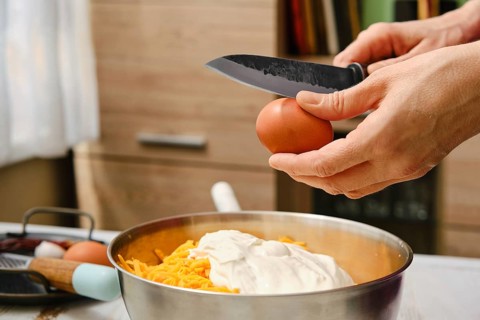 A person cracks an egg over a bowl filled with grated cheese and sour cream. The setting is a