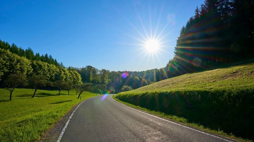 A winding country road under clear skies and bright sun, surrounded by green meadows and forests,