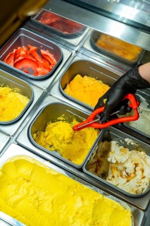 Person wearing black glove scooping a vibrant yellow gelato from a metal tray in a brightly lit