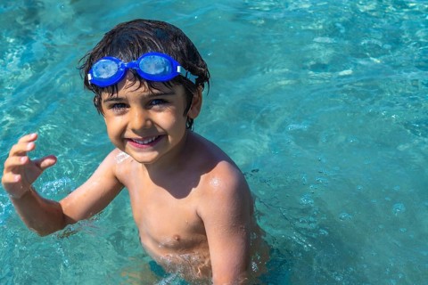 Young boy with blue swim goggles smiling at camera while swimming and playing in the clear,