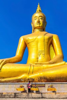 Monumental golden Buddha statue against deep blue sky, visitor and offerings at the pedestal,