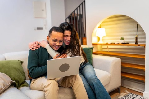 Cheerful multiethnic couple cuddling on a modern sofa, smiling and watching a movie or browsing on